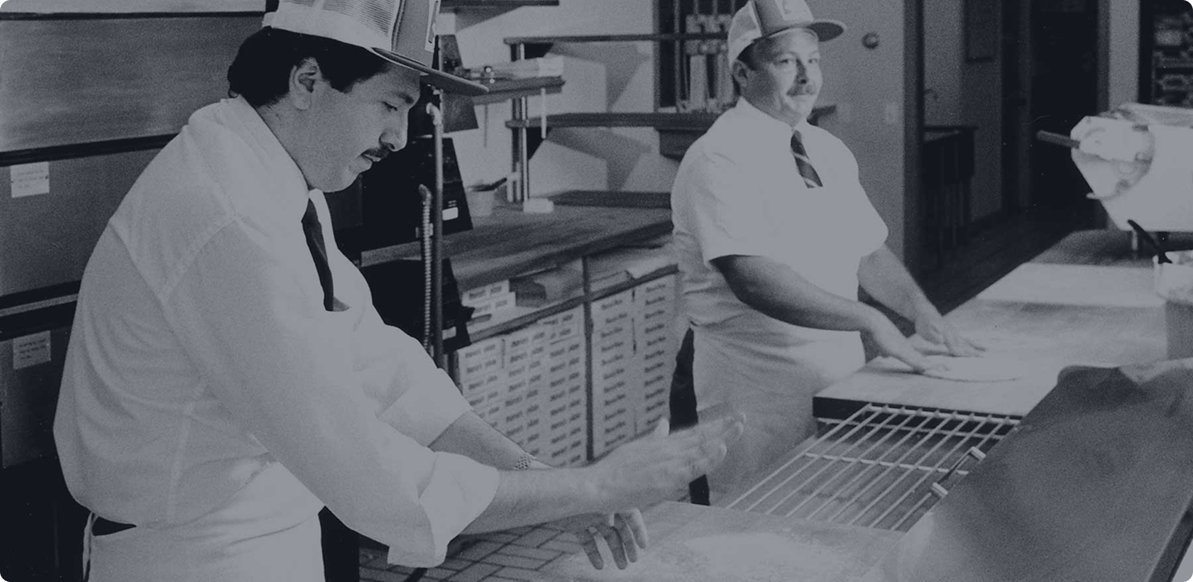 Two men in a kitchen rolling out pizza dough